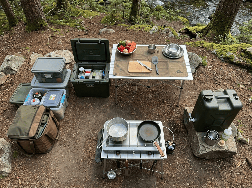 Outdoor cooking station with stove, cookware, food containers, and cooler beside a forest stream